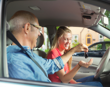 Smiling female driver holding the car steering wheel while the driving instructor sitting next to her is handing over car keys